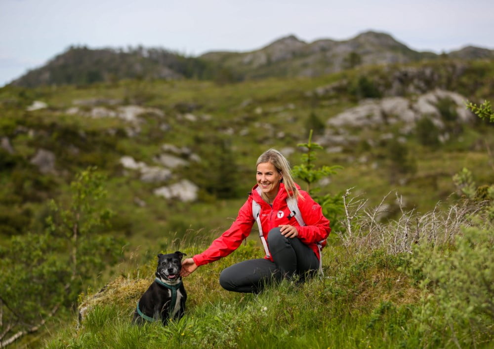 En dame i rød turjakke fra Turistforeningen sitter i fjellheimen og koser med en hund