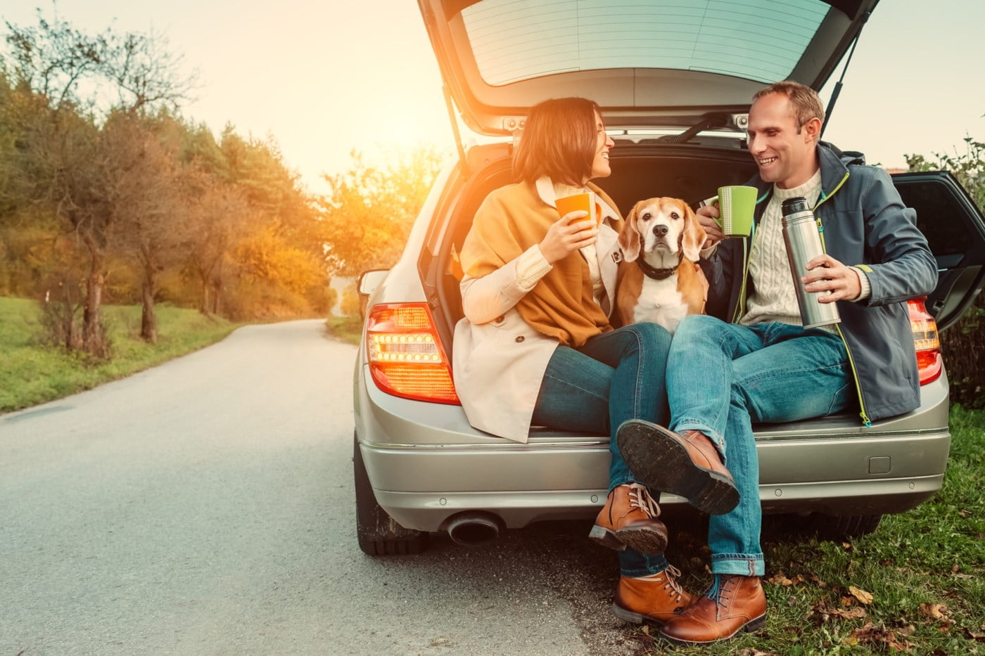 En kvinne, en mann og en hund sitter i bagasjerommet til en bil og tar en kaffepause.